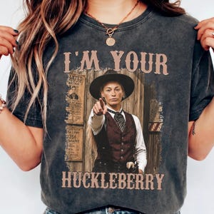 May include: A charcoal gray t-shirt featuring a graphic print of a person in a cowboy hat and vest pointing forward. The text "I'M YOUR HUCKLEBERRY" is printed in a western-style font above and below the image, alongside a vintage wanted poster.