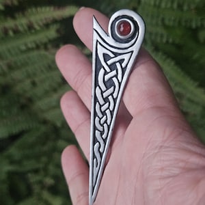 May include: A silver-toned metal brooch featuring an elongated triangular shape with an intricate embossed Celtic knotwork pattern. The top of the piece is rounded and contains a small, circular, deep red stone set into the center of the metal.