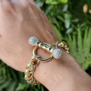May include: A gold-toned chain link bracelet featuring a toggle clasp closure. The toggle bar is accented with two spherical ends covered in small, clear, reflective stones that provide a sparkling appearance against the polished metal links.