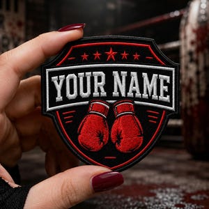 Può includere: Una toppa ricamata a forma di scudo con sfondo nero e bordo rosso. Il design include due guantoni da boxe rossi, cinque stelle rosse nella parte superiore e il testo bianco "YOUR NAME" centrato orizzontalmente nella sezione centrale.