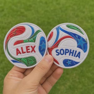 May include: Two circular embroidered patches featuring a soccer ball pattern in red, blue, and green thread. The left patch displays the name ALEX in red, while the right patch displays the name SOPHIA in blue, both set against a white background.