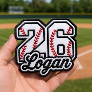May include: A custom embroidered patch featuring the number 26 in white with red baseball-style stitching details, outlined in black. Below the number, the name Logan is embroidered in a black cursive script font with a white border.