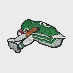 May include: An embroidered patch featuring a football player in a green jersey with the number 2, white trousers, and a brown football tucked under an arm. The player is depicted in a dynamic, forward-leaning pose with black outlines and stitching.