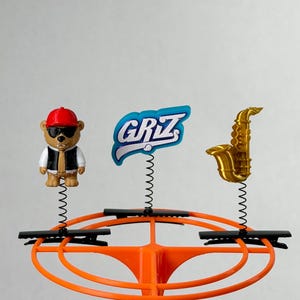May include: Three decorative toppers attached to black hair clips on an orange circular stand. The toppers include a bear wearing a red cap and sunglasses, a blue and white sign with the text GRIZ, and a gold-coloured saxophone figurine.