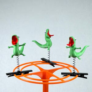 Könnte beinhalten: Drei grüne Echsenfiguren mit geöffneten Mäulern und roten Zungen, die jeweils auf einer dünnen schwarzen Metallfeder montiert sind. Diese Federn sind an schwarzen Krokodilklemmen befestigt, die am oberen Rand eines runden orangefarbenen Kunststoffständers sitzen.