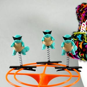 Puede incluir: Tres figuras de color azul claro con forma de tortuga, caparazones beige y gafas de sol negras, montadas sobre un muelle metálico negro unido a una pinza de cocodrilo negra, sobre un soporte circular de plástico naranja.
