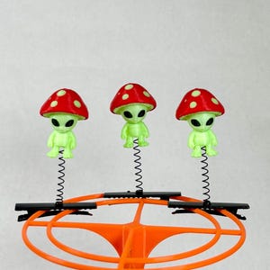May include: Three light green alien figures with large black eyes, each wearing a red mushroom cap with white spots. Each figure is attached to a thin black metal spring mounted on a black alligator clip, all secured to an orange plastic frame.