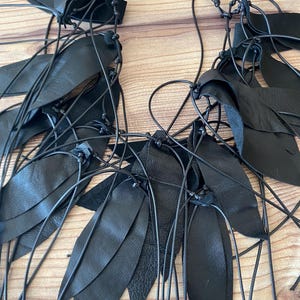 Puede incluir: Un collar negro que presenta numerosas piezas de cuero en forma de hoja unidas a finos cordones oscuros. El dise&ntilde;o consiste en m&uacute;ltiples segmentos de cuero negro superpuestos y atados con nudos, creando una disposici&oacute;n en cascada de follaje oscuro.