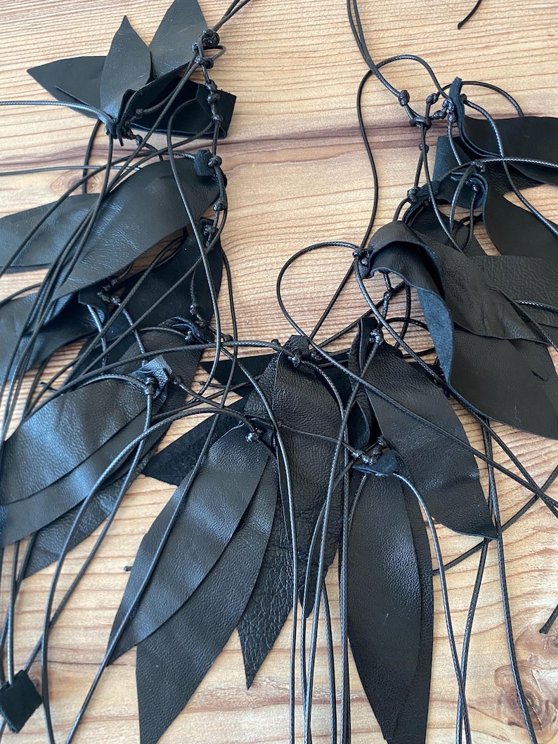 Puede incluir: Un collar negro que presenta numerosas piezas de cuero en forma de hoja unidas a finos cordones oscuros. El dise&ntilde;o consiste en m&uacute;ltiples segmentos de cuero negro superpuestos y atados con nudos, creando una disposici&oacute;n en cascada de follaje oscuro.