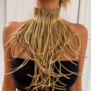 May include: A gold-coloured statement necklace featuring a high collar design composed of numerous thin, flexible, metallic-looking strands that drape downwards over the chest, creating a fringe-like effect that covers the upper torso area.
