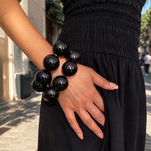 Könnte beinhalten: Ein Armband bestehend aus mehreren großen, glänzend schwarzen, kugelförmigen Perlen, die in einer Gruppe angeordnet sind. Die Perlen haben eine glatte, reflektierende Oberfläche und sind zu einer runden, massiven Form verbunden.
