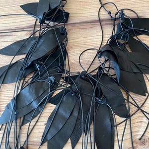Puede incluir: Un collar negro que presenta m&uacute;ltiples recortes de cuero en forma de hoja unidos a cordones negros finos y anudados. El dise&ntilde;o consiste en piezas de cuero puntiagudas y superpuestas dispuestas en grupos a lo largo de la estructura de cordones.
