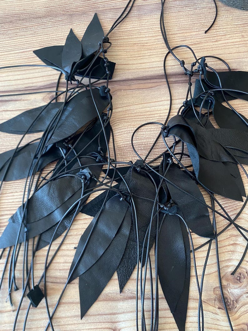 Puede incluir: Un collar negro que presenta m&uacute;ltiples recortes de cuero en forma de hoja unidos a cordones negros finos y anudados. El dise&ntilde;o consiste en piezas de cuero puntiagudas y superpuestas dispuestas en grupos a lo largo de la estructura de cordones.