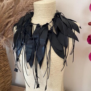 Puede incluir: Un collar negro que presenta m&uacute;ltiples recortes de cuero en forma de hoja unidos a finos cordones de cuero negro. El dise&ntilde;o consiste en piezas de cuero negro puntiagudas y superpuestas que caen alrededor del cuello, aseguradas con nudos.
