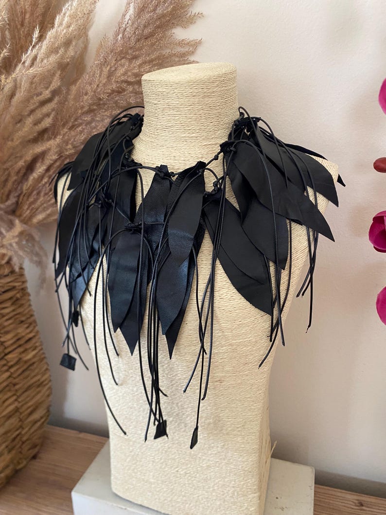 Puede incluir: Un collar negro que presenta m&uacute;ltiples recortes de cuero en forma de hoja unidos a finos cordones de cuero negro. El dise&ntilde;o consiste en piezas de cuero negro puntiagudas y superpuestas que caen alrededor del cuello, aseguradas con nudos.