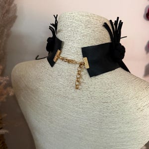 May include: A black choker necklace featuring a wide band with a textured surface. The ends are connected by a gold-toned metal chain with a lobster clasp and an adjustable extender. Black fabric tassels and circular accents are attached to the sides.