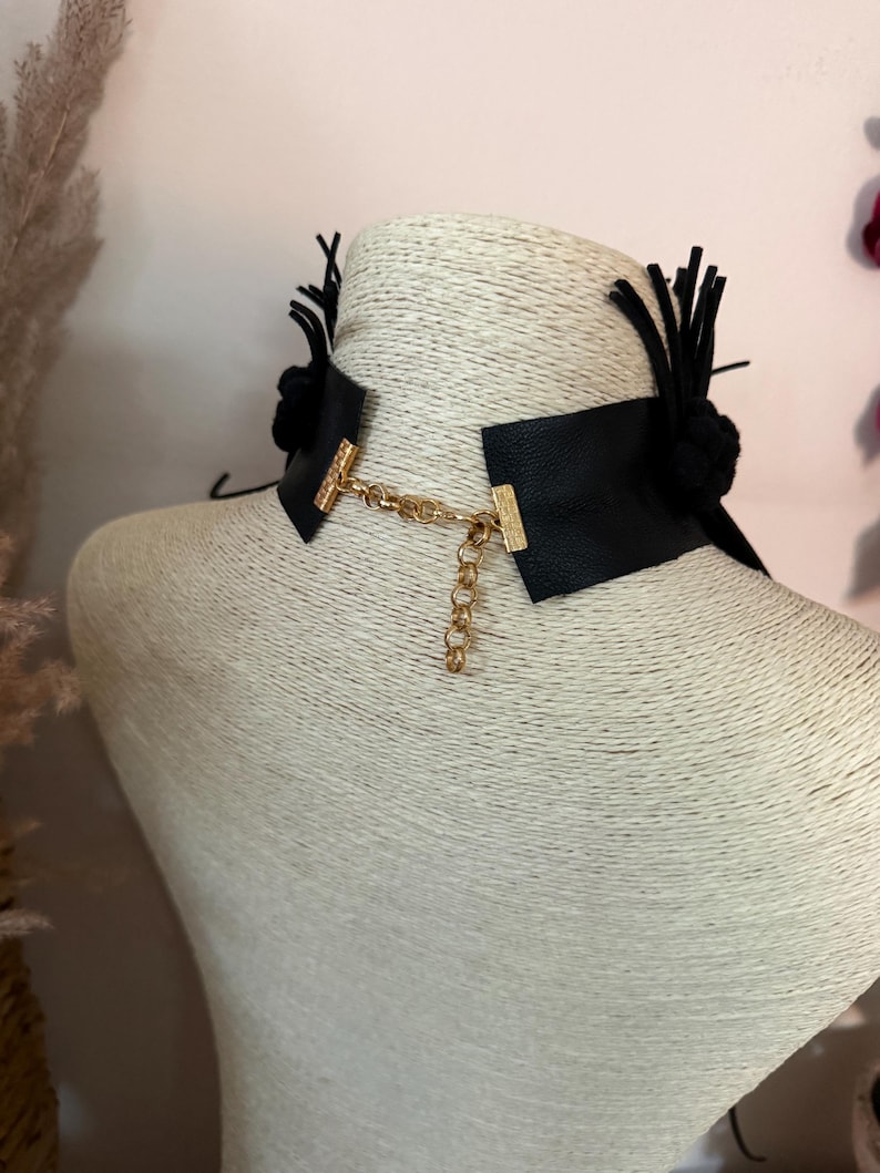 May include: A black choker necklace featuring a wide band with a textured surface. The ends are connected by a gold-toned metal chain with a lobster clasp and an adjustable extender. Black fabric tassels and circular accents are attached to the sides.