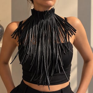 Puede incluir: Un collar tipo gargantilla negro con un diseño de cuello alto y estructurado, con largas y finas tiras de flecos colgantes que se extienden sobre el pecho. El accesorio está construido con múltiples segmentos verticales, creando una apariencia texturizada.