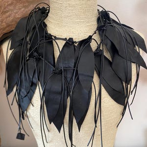 Puede incluir: Un collar negro llamativo construido con m&uacute;ltiples piezas de cuero en forma de hoja unidas a finos cordones negros. El dise&ntilde;o presenta segmentos puntiagudos y superpuestos que caen alrededor del cuello, creando una apariencia texturizada con flecos.