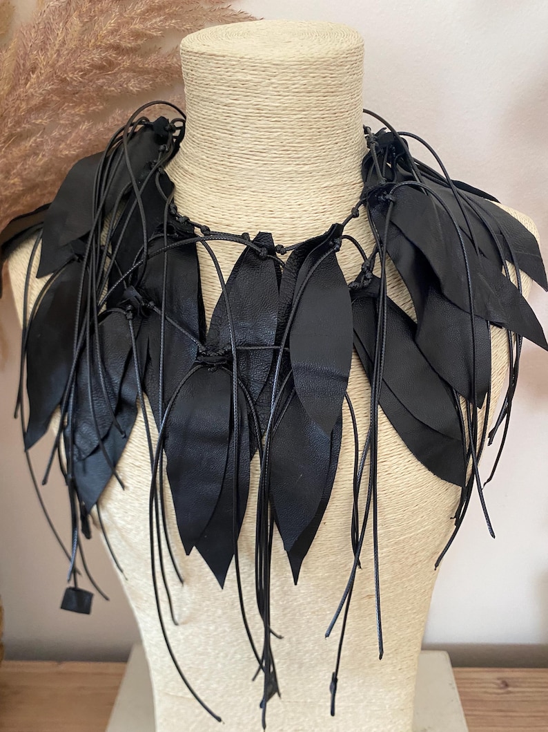 Puede incluir: Un collar negro llamativo construido con m&uacute;ltiples piezas de cuero en forma de hoja unidas a finos cordones negros. El dise&ntilde;o presenta segmentos puntiagudos y superpuestos que caen alrededor del cuello, creando una apariencia texturizada con flecos.