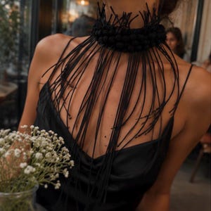 Puede incluir: Un collar tipo gargantilla negro con una banda gruesa y texturizada adornada con numerosos cordones negros largos, finos y colgantes que caen sobre el pecho. El accesorio se combina con una blusa negra sin mangas con escote drapeado.