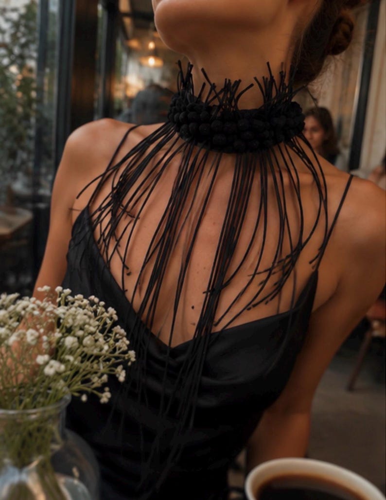 May include: A black choker necklace featuring a thick, textured band adorned with numerous long, thin, dangling black cords that drape over the chest. The accessory is paired with a simple black sleeveless top with a draped neckline.