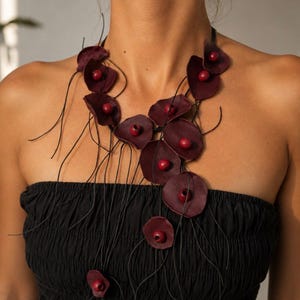 Puede incluir: Un collar con múltiples elementos en forma de pétalo de color burdeos oscuro dispuestos a lo largo de finos cordones negros. Cada pieza en forma de pétalo está acentuada con una pequeña cuenta redonda de color burdeos a juego en su centro.