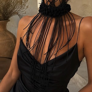 May include: A black choker necklace featuring a thick, textured band adorned with numerous long, thin, dangling black cords that drape over the chest. The accessory is worn with a black satin slip dress with thin spaghetti straps and a V-neckline.