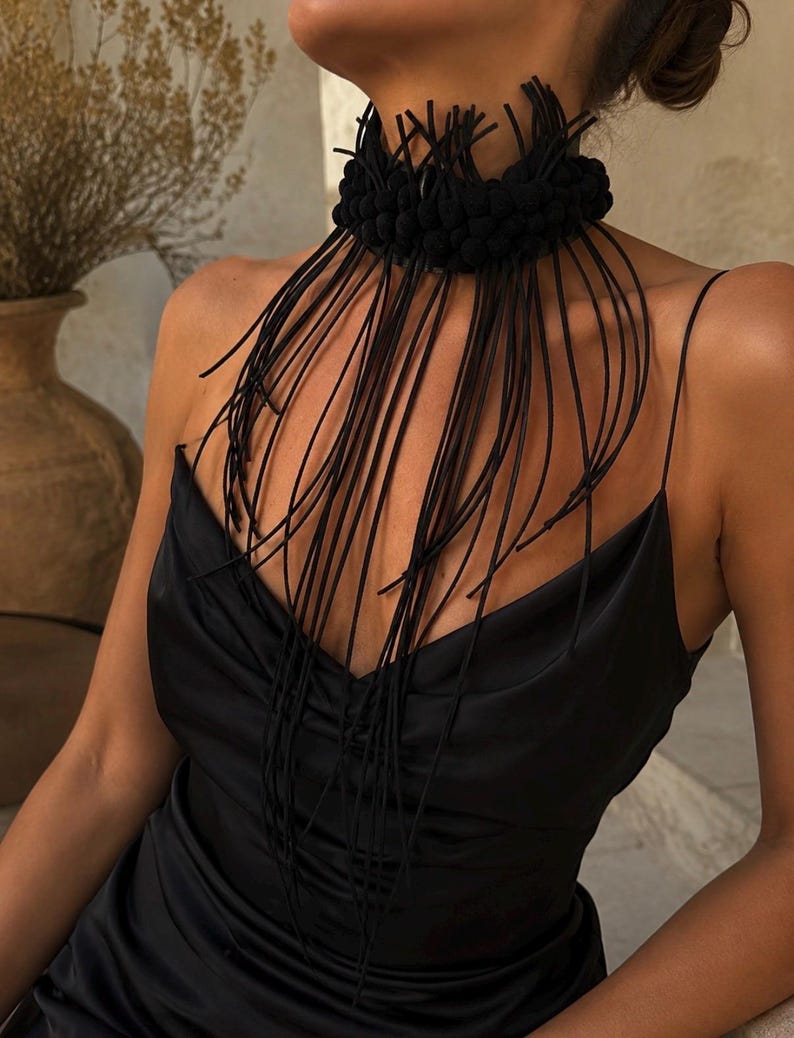 May include: A black choker necklace featuring a thick, textured band adorned with numerous long, thin, dangling black cords that drape over the chest. The accessory is worn with a black satin slip dress with thin spaghetti straps and a V-neckline.