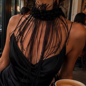 May include: A black choker necklace featuring a textured, beaded collar base with long, thin, dangling black fringe strands that drape over the chest, paired with a black satin slip dress with thin spaghetti straps and a V-neckline.