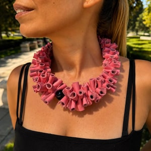 Puede incluir: Un collar llamativo que presenta numerosos tubos pequeños enrollados de color rosa dispuestos en un patrón circular denso y con volantes. Una cuenta negra única, lisa y redonda, está integrada en la parte frontal del collar.