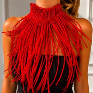 Puede incluir: Un collar de flecos de color rojo vibrante que presenta numerosos hilos largos y delgados similares a cordones que caen desde una banda de cuello alta y estructurada. El accesorio se lleva sobre una prenda negra sin tirantes.