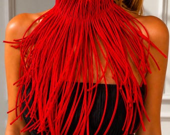 May include: A vibrant red fringe collar necklace featuring numerous long, thin, cord-like strands that drape from a high, structured neckband. The accessory is worn over a black, textured, strapless garment, creating a bold contrast in color.