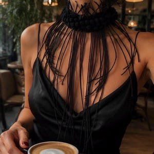 May include: A black choker necklace featuring a thick, textured band adorned with numerous small, spherical beads and long, thin, dangling black fringe strands that drape over the chest, paired with a black satin camisole with thin spaghetti straps.