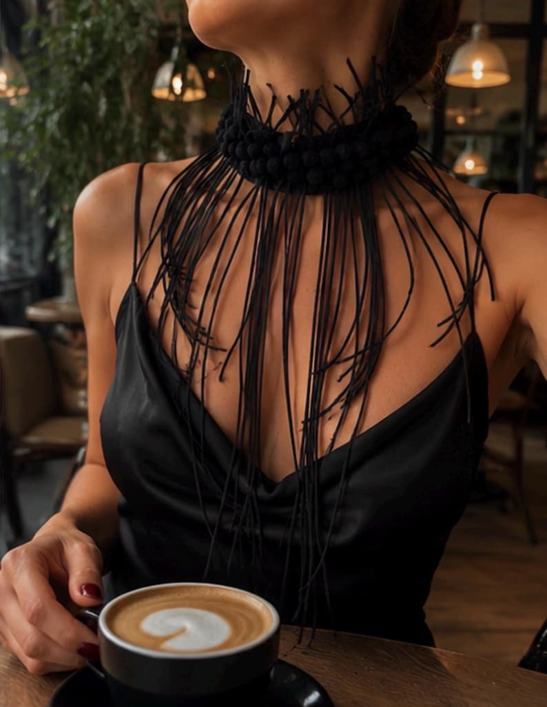 May include: A black choker necklace featuring a thick, textured band adorned with numerous small, spherical beads and long, thin, dangling black fringe strands that drape over the chest, paired with a black satin camisole with thin spaghetti straps.