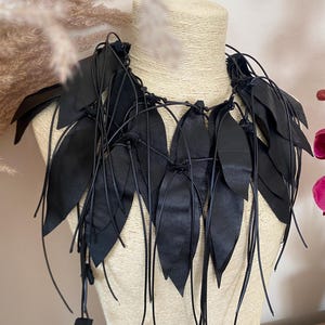 Puede incluir: Un collar de cuero negro con m&uacute;ltiples recortes en forma de hoja unidos a finos cordones negros. El dise&ntilde;o consiste en piezas de cuero puntiagudas y superpuestas que cuelgan alrededor del cuello, aseguradas con nudos y extremos de cord&oacute;n largos.