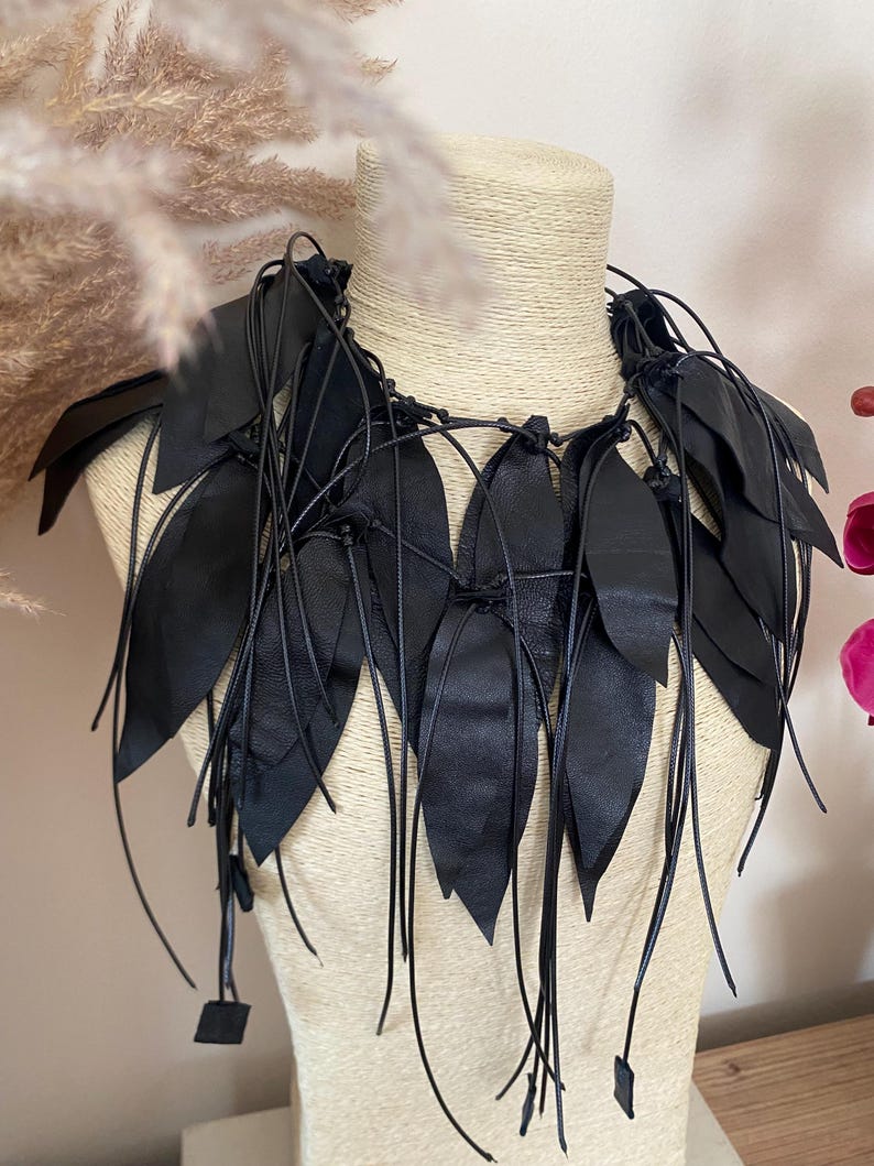 Puede incluir: Un collar de cuero negro con m&uacute;ltiples recortes en forma de hoja unidos a finos cordones negros. El dise&ntilde;o consiste en piezas de cuero puntiagudas y superpuestas que cuelgan alrededor del cuello, aseguradas con nudos y extremos de cord&oacute;n largos.