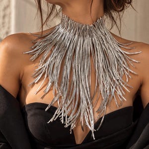Puede incluir: Un collar plateado llamativo con un diseño de cuello alto compuesto por numerosos hilos metálicos finos y texturizados que caen en un patrón de flecos, creando un efecto denso y superpuesto alrededor del cuello y la parte superior del pecho.