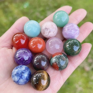 Op de afbeelding: Een verzameling van twaalf kleine, gepolijste, bolvormige edelstenen in diverse kleuren, waaronder rood, blauw, groen, paars, roze en bruin. Elke steen heeft een glad, reflecterend oppervlak met duidelijke natuurlijke patronen en texturen.