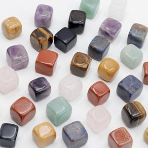 May include: A collection of small, polished stone cubes in various colours and textures, including purple amethyst, orange tiger eye, red jasper, black obsidian, light green aventurine, pale pink rose quartz, and dark blue sodalite, arranged in a grid.