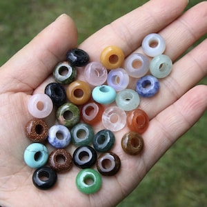 Può includere: Una collezione di piccole perline rotonde a forma di ciambella in vari colori e texture, tra cui finiture traslucide, opache, maculate e solide. Le perline sono realizzate in diversi minerali e pietre, con tonalità di blu, verde, marrone, nero, rosa e bianco.