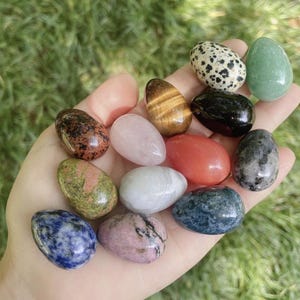 Puede incluir: Una colección de trece piedras pequeñas, lisas, pulidas y con forma de huevo en varios colores y patrones, incluyendo jaspe dálmata moteado, ojo de tigre, cuarzo rosa, sodalita, unakita y otros especímenes minerales variados.