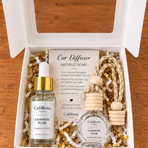 May include: A white gift box containing a 15 mL glass bottle of CaliRene Candles Cashmere Plum fragrance refill with a gold dropper, a glass car diffuser with a wooden cap and hanging cord, and an instruction card for the car diffuser.