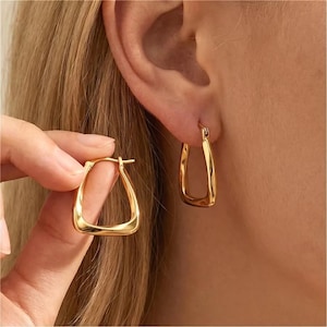 May include: A pair of gold-toned hoop earrings with a distinct triangular or U-shaped silhouette. One earring is worn in the earlobe, while the other is held between fingers, showcasing the smooth, polished metallic surface and the hinged clasp mechanism.