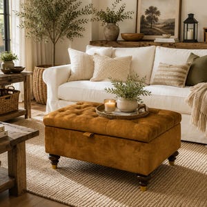 May include: A rectangular, tufted storage ottoman upholstered in a mustard-brown fabric. The ottoman features a button-tufted top, a hinged lid with a small pull tab, and four dark wooden legs with gold-toned feet, resting on a woven rug.