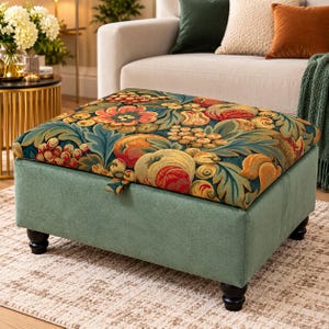 May include: A square storage ottoman with a solid sage green fabric base and four dark brown turned wooden feet. The hinged lid features a vibrant tapestry-style fabric with a dense pattern of colourful fruits, berries, and green leaves.