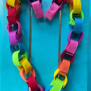 May include: A rainbow felt chain in the shape of a heart. The chain is made of individual felt circles linked together. The colors of the chain are red, orange, yellow, green, blue, indigo, and violet.