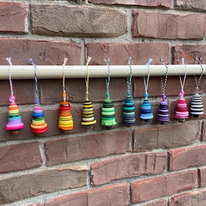 May include: A row of 12 colorful Christmas tree ornaments made from stacked buttons. The ornaments are hanging from a white rod against a brick wall.