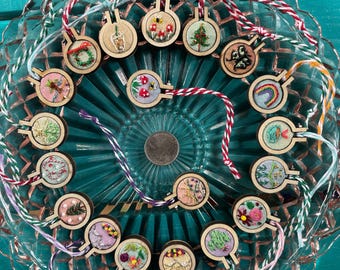 May include: A collection of small, round wooden embroidery hoops with various colorful embroidered designs, including plants, insects, and rainbows. Each hoop has a loop of string for hanging. The hoops are arranged on a clear, decorative glass plate.