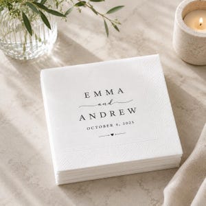 May include: A stack of square white paper napkins featuring black printed text that reads EMMA and ANDREW, followed by the date OCTOBER 4, 2025, and a small heart icon centred between two horizontal lines at the bottom of the design.